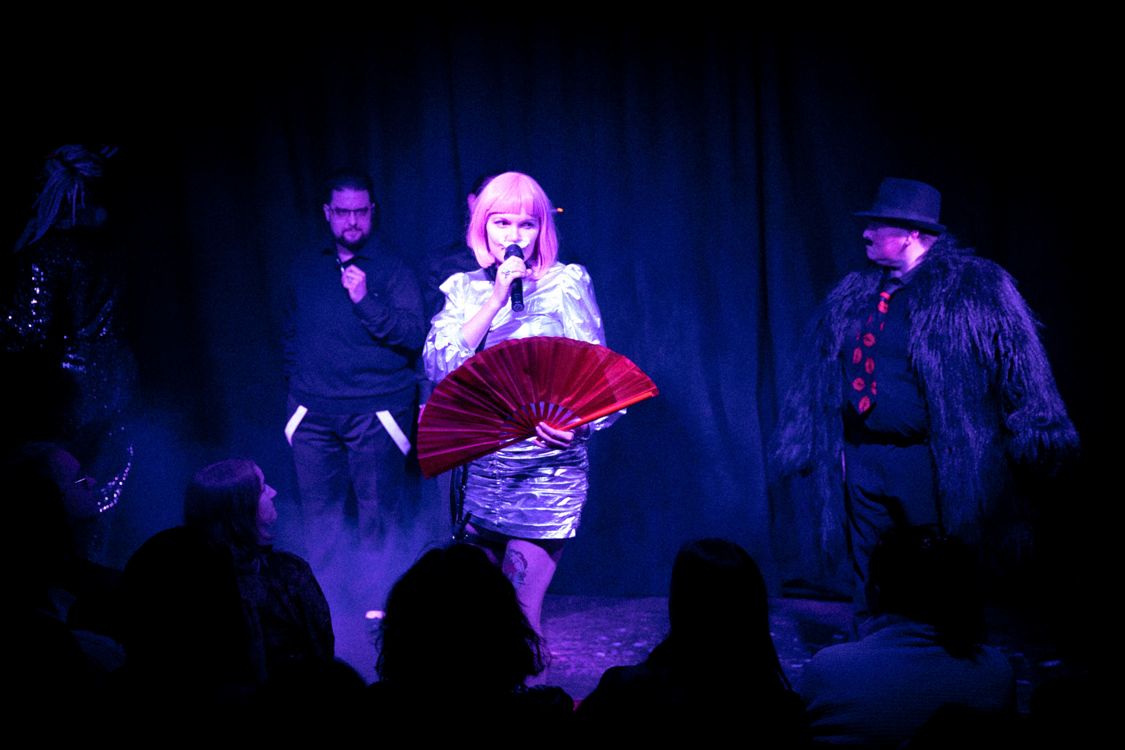 Na scenie występuje oświetlona reflektorem drag queen w różowej perółce, z białymi wąsami, trzyma czerwony wachlarz, na tle kurtyny widoczne są osoby w monochromatycznych czarnych kostiumach.