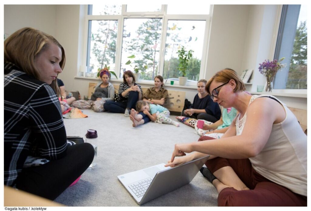 Grupa kobiet w kręgu, na podłodze. Na pierwszym planie osoba pokazuje coś na laptopie pozostałym uczestniczkom