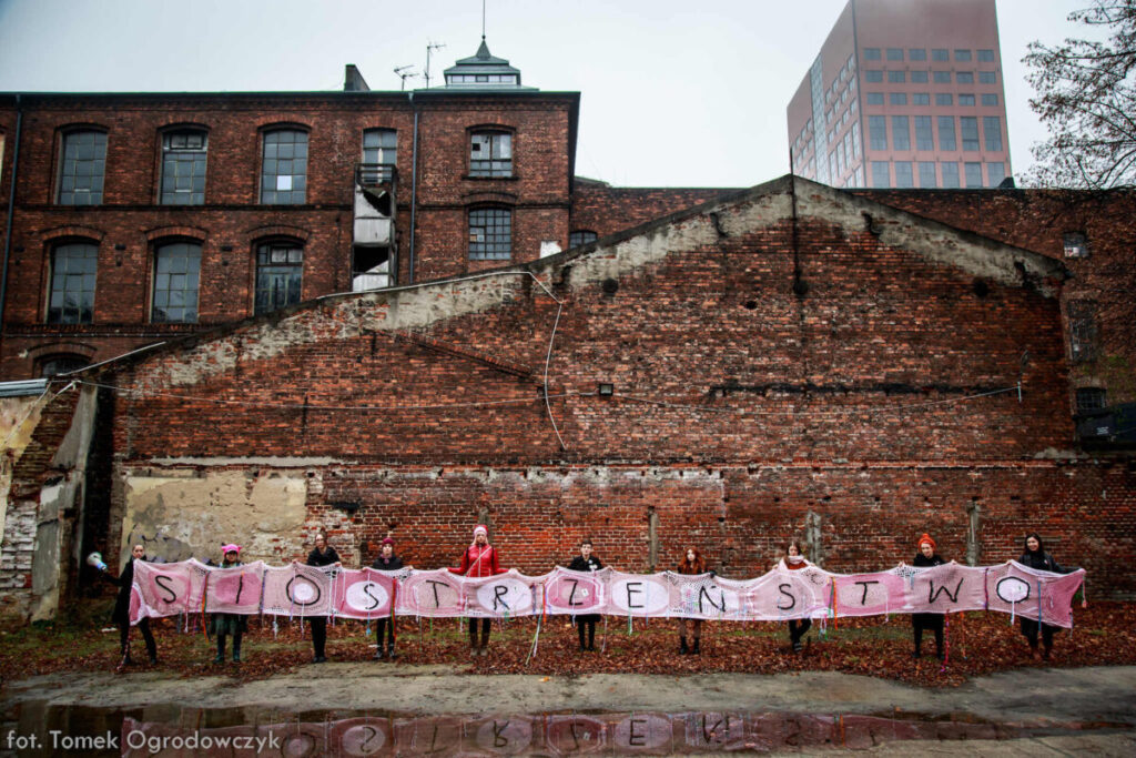 Dziesięć osób trzymających duży, biało-różowy transparent z napisem „Siostrzeństwo” zrobiony na drutach, w tle stare budynki z czerwonej cegły