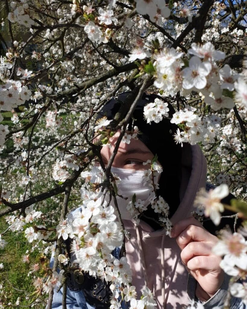 Dziewczyna w maseczce na twarzy i chuście na głowie spoglądająca spomiędzy gałęzi kwitnącego drzewa