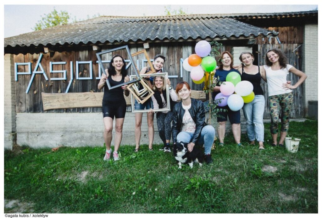 7 kobiet i pies na tle drewnianego budynku z napisem "Pasjodzielnia". Osoby śmieją się, trzymają balony i ramki na zdjęcia