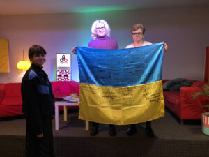 Trzy osoby, w tym dwie starsze kobiety trzymają flagę Ukrainy z podpisami wsparcia od ludzi.