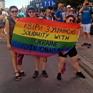 Trzy osoby trzymają tęczową flagę z napisem 'Solidarnie z Ukrainą' w języku ukraińskim i angielskim podczas protestu.